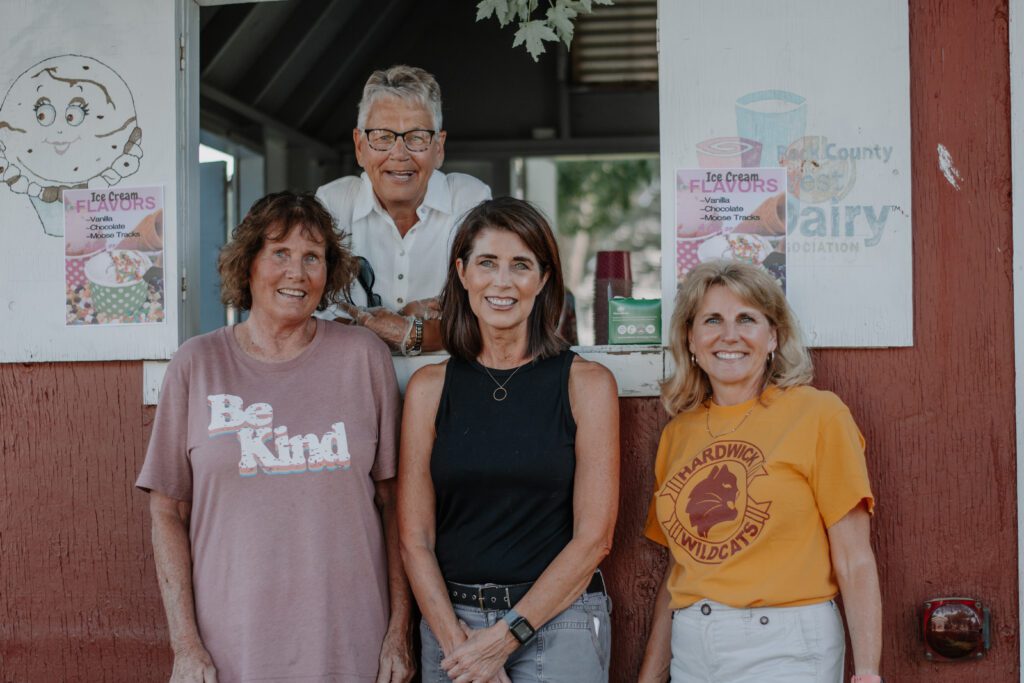 Four women stand together to take a group photo in front of a wooden ice cream stand, with one leaning out the serving window.