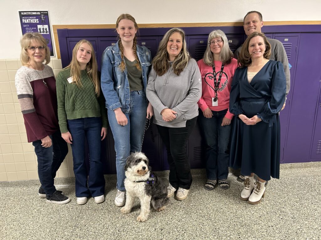 A group photo with Phoebe the Aussiedoodle in the hallway at Glencoe-Silver Lake Elementary School.