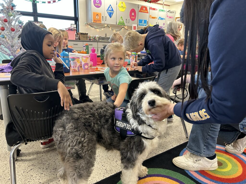 Phoebe the Aussiedoodle looks at the camera while two young students and a teacher pet her.