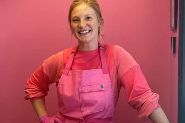 Kenzie wears a pink apron over a pink sweatshirt and stands in front of a pink wall in her small business, Pink Apron Bakery.