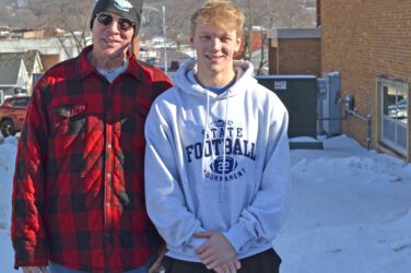 Two people stand outside in the snow, smiling. One wears a red plaid jacket, sunglasses, and a knit hat, while the other wears a light gray Football State Tournament hoodie. A brick building and houses are in the background.