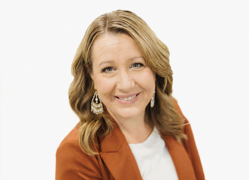 A woman with wavy, shoulder-length blonde hair smiles at the camera. She wears a rust-colored blazer, white top, and large, dangling earrings, set against a plain white background.