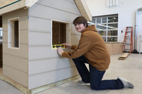 A student kneels on the shop floor at JCC and uses a level in the window of a small shed being built.