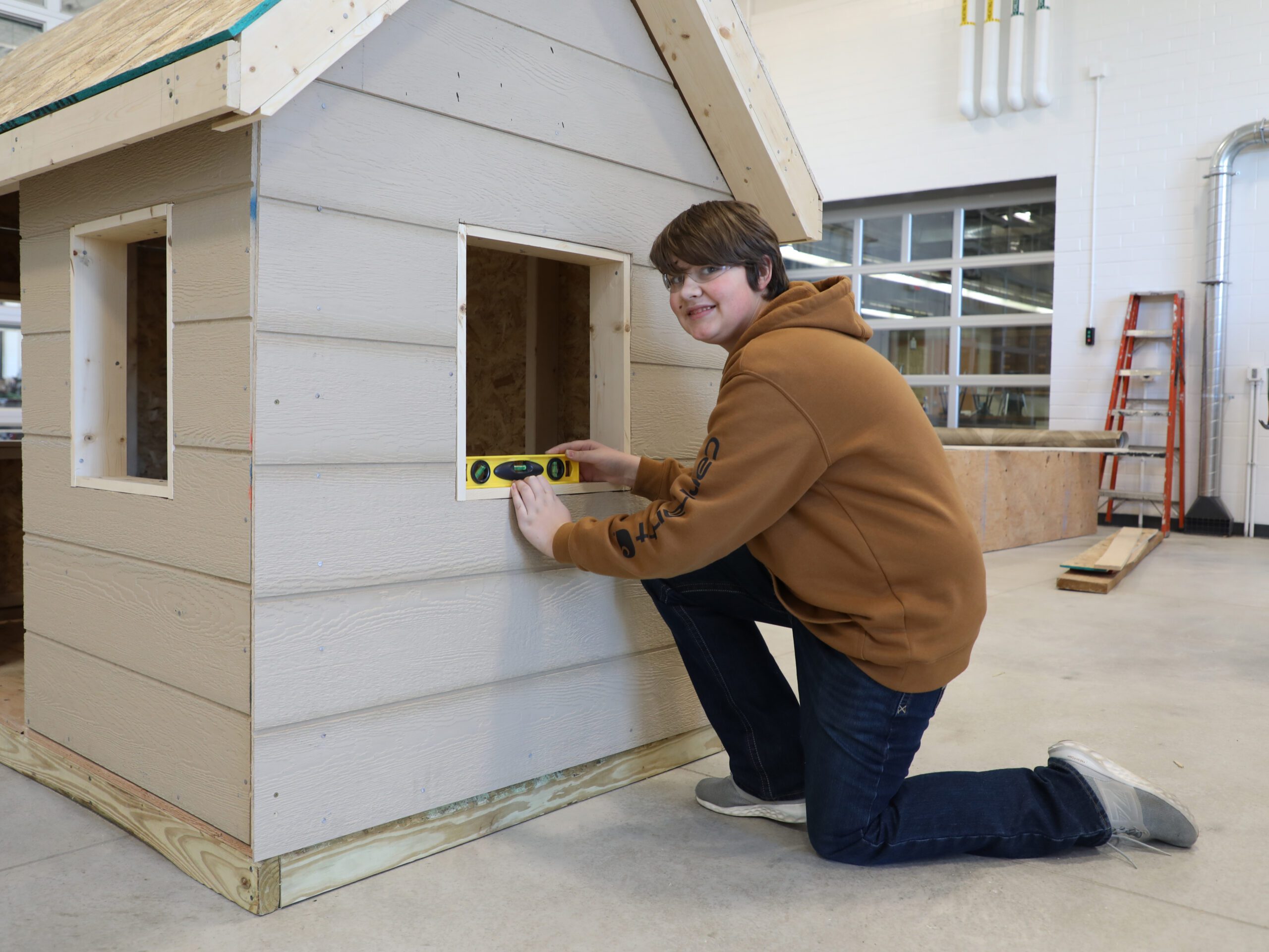 A student kneels on the shop floor at JCC and uses a level in the window of a small shed being built.