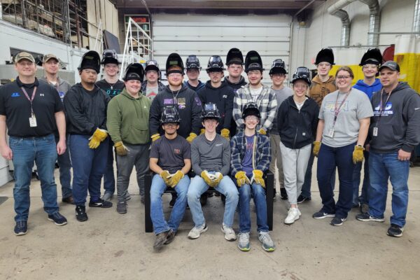 A group of students wearing welding safety helmets pose for a photo with educators and mentors.