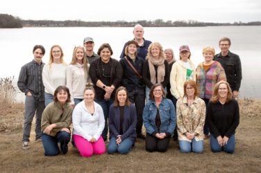 A group of seventeen adults pose together on grass in front of a lake on an overcast day. Some are standing, and others are kneeling or sitting, all smiling at the camera. Trees and water are visible in the background.