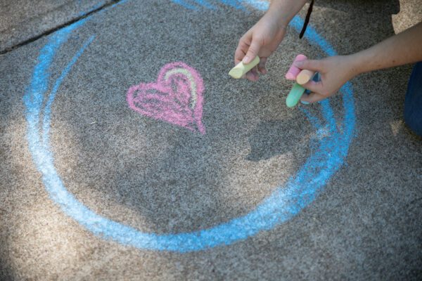 A person's hands hold four colors of chalk above a drawing of a heart in a circle on the sidewalk.