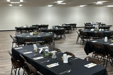 A banquet hall with several tables covered in black tablecloths, each set with plates, utensils, cups, and simple centerpieces. Brown chairs surround the tables, and the room has a wood floor and white walls.