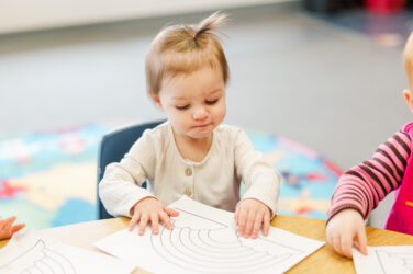A young toddler with light brown hair sits at a table, touching a piece of paper with a rainbow outline. The child is focused, reminding us of the many ways to appreciate child care providers who nurture such creativity. A colorful rug is visible in the background.
