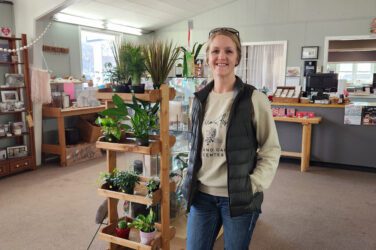 A woman wearing a vest and sunglasses on her head stands smiling next to a display of potted plants inside a bright, cozy shop with shelves, a counter, and various decor items.