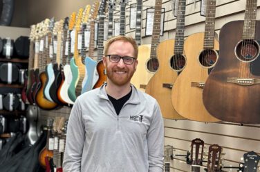 A smiling man with glasses and a beard stands in front of a wall display of acoustic and electric guitars in a music store. He is wearing a light gray pullover with a logo on the left chest.