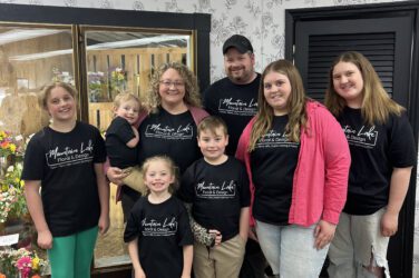 A group of seven people, including children and adults, all wearing matching Mountain Lake Floral shirts, pose and smile together in a flower shop surrounded by beautiful flowers and creative floral arrangements.