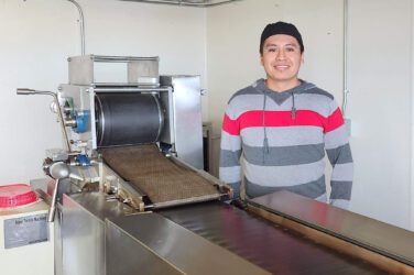A person wearing a striped hoodie and black cap stands next to a tortilla making machine at Tortilleria El Maná in a kitchen or food production area, smiling at the camera.