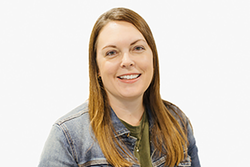 A woman with straight, light brown hair wearing a denim jacket over a green top, smiling at the camera against a plain white background.