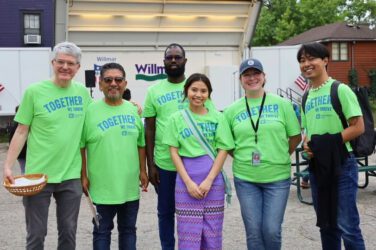 Six people smiling and standing together outdoors, all wearing matching bright green Together We Thrive T-shirts, with buildings and trees visible in the background.