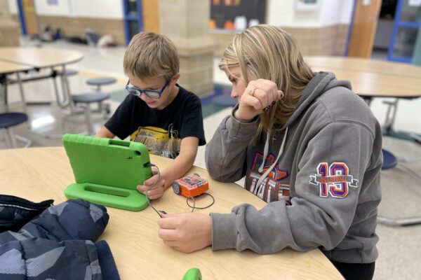 An adult helps a child using a bright green tablet, both sitting at a round wooden table.