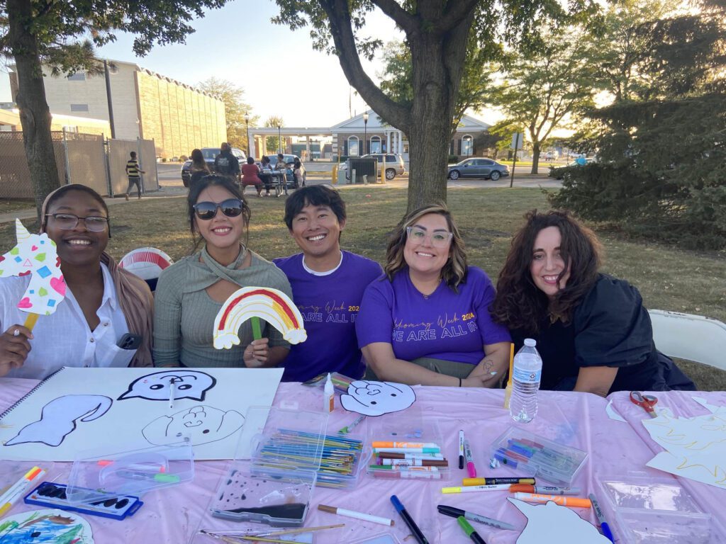 Suree sits in a group at a craft table and holds up a paper rainbow during Welcoming Week in Worthington.