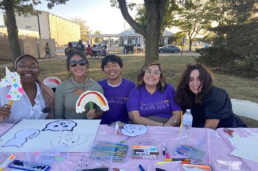 Suree sits in a group at a craft table and holds up a paper rainbow during Welcoming Week in Worthington.