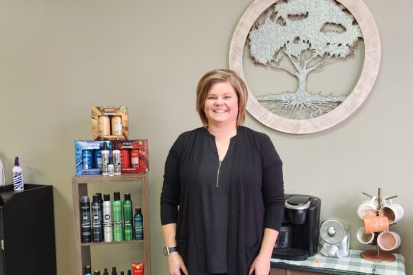 Kirsten stands inside her salon with a large wood tree cutout on the wall behind her and a display of hair products next to her.