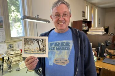 A smiling man wearing glasses and a blue Free Ice Water Wall Drug T-shirt stands in an office, holding up a postcard with an illustrated outdoor scene. A desk with papers and office supplies is visible behind him.