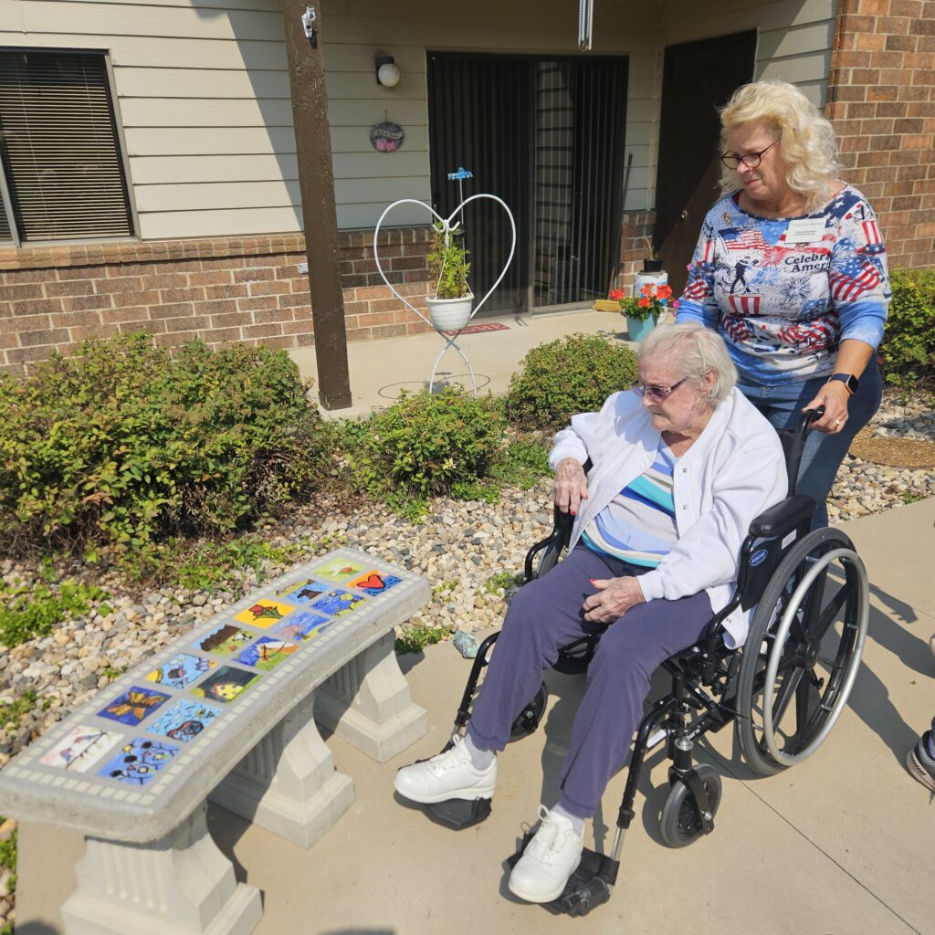 A resident in a wheel chair checks out the new concrete bench with art tiles, helped by a staff member.