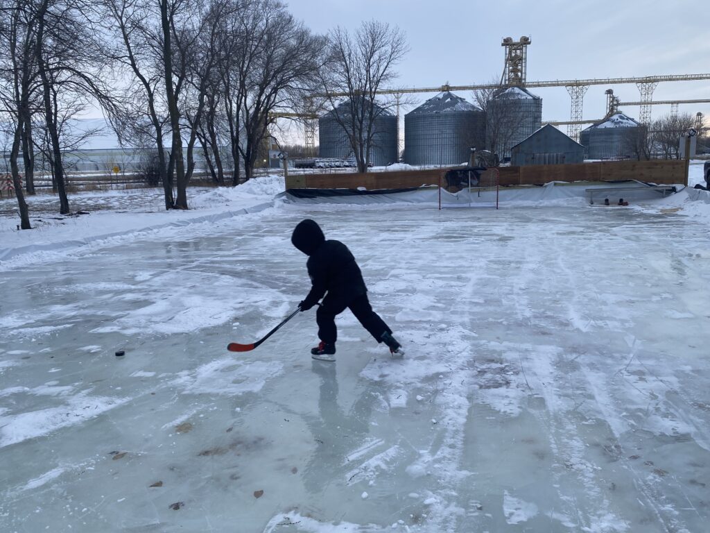 A child brandishing a hockey stick skates on an outdoor rink with grain bins in the distance.