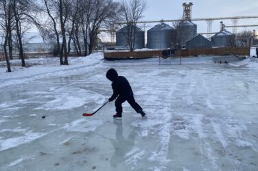 A child brandishing a hockey stick skates on an outdoor rink with grain bins in the distance.