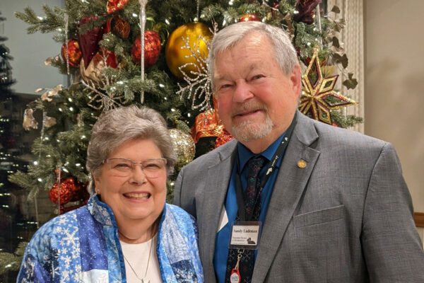 Peggy and Sandy Ludeman smile in front of a decorated Christmas tree. The woman wears a blue patterned jacket and glasses; the man wears a gray suit and name badge. The tree is adorned with red, gold, and white ornaments.