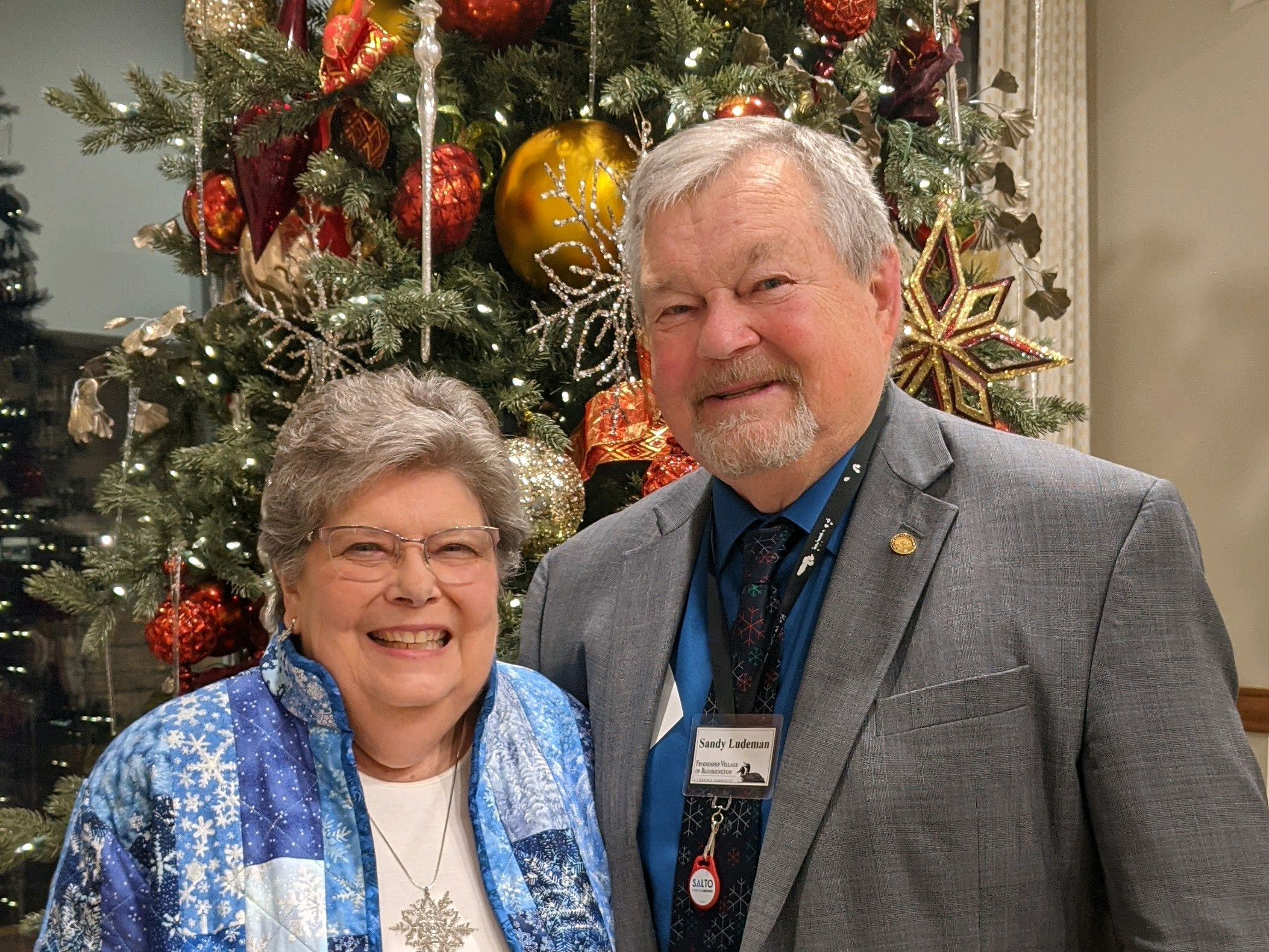 Peggy and Sandy Ludeman smile in front of a decorated Christmas tree. The woman wears a blue patterned jacket and glasses; the man wears a gray suit and name badge. The tree is adorned with red, gold, and white ornaments.