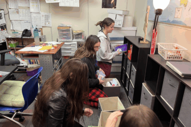 Four people are organizing and sorting items into black storage cubbies and bins in a classroom or office space filled with shelves, papers, and various supplies.