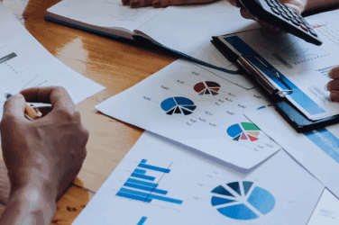 Close-up of people’s hands working with financial charts, graphs, and documents on a wooden table, using a calculator and a pen, suggesting a business meeting or data analysis discussion.