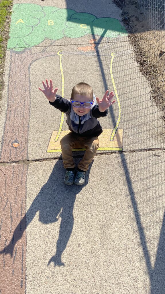 A young child raises their hands while sitting on the picture of a swing painted on the sidewalk as part of the Born Learning Trail in Madison.