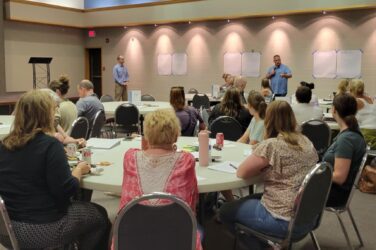 A group of people sit at round tables in a conference room, listening to two men standing at the front near blank sheets on the wall. Papers, drinks, and food are on the tables. The setting appears to be a workshop or seminar.