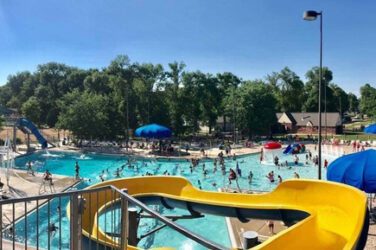 The pool is full of people on a summer day, with a yellow slide in the foreground.