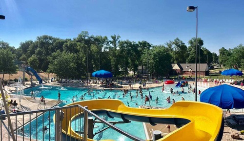 The pool is full of people on a summer day, with a yellow slide in the foreground.