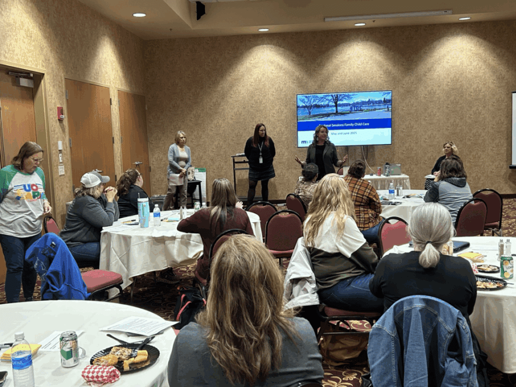 Presenters stand by a PowerPoint displayed at the front of a room of people seated around tables in a child care listening session.