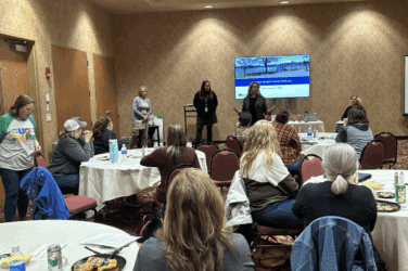 Presenters stand by a PowerPoint displayed at the front of a room of people seated around tables in a child care listening session.