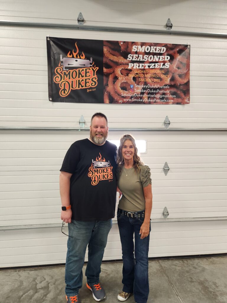 Lance and Mari stand under a banner with "Smokey Dukes" and an image of pretzels on it.