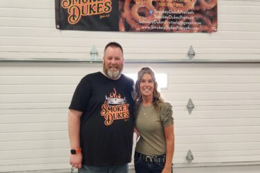 Lance and Mari stand under a banner with "Smokey Dukes" and an image of pretzels on it.