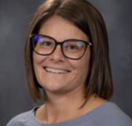 A woman with straight brown hair and glasses smiles at the camera. She is wearing a gray top and is posed in front of a dark, blurred background.