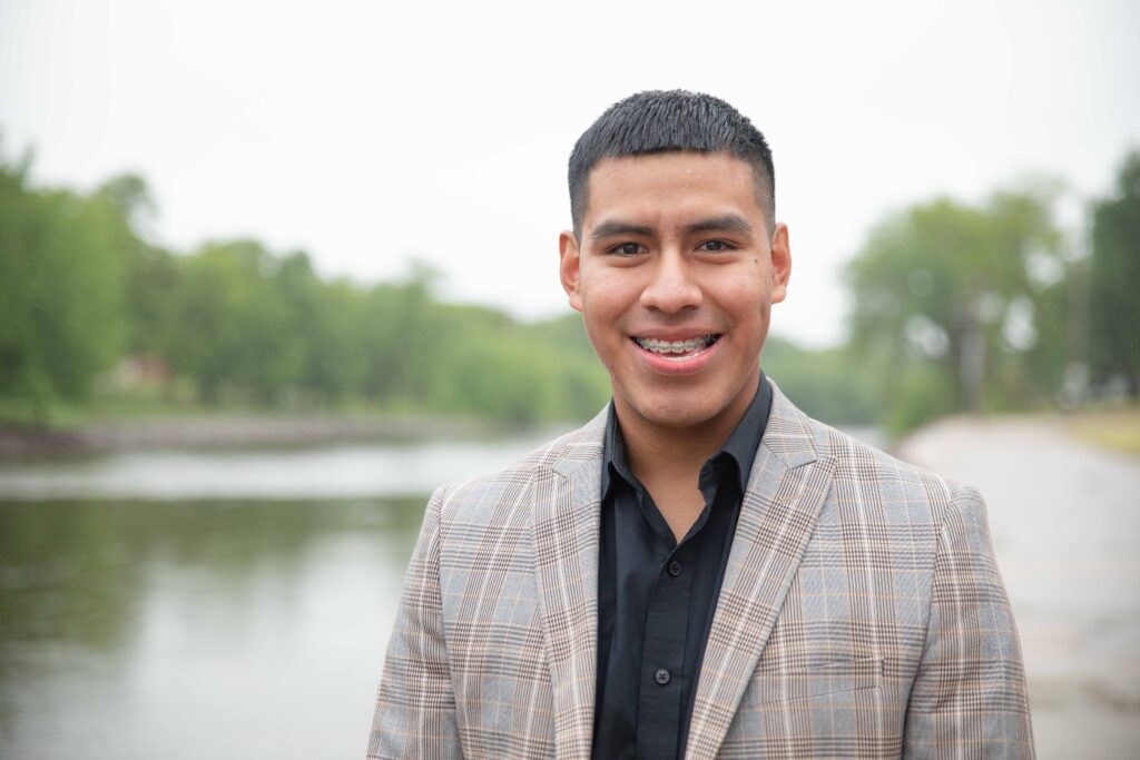 Alex poses for a head shot om a plaid blazer and black shirt, with a river and trees in the background.