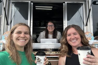 Marie and Meghan hold up cups of coffee from Heather, who stands inside her mobile coffee trailer, Mocha Motion