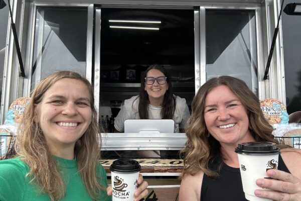 Marie and Meghan hold up cups of coffee from Heather, who stands inside her mobile coffee trailer, Mocha Motion