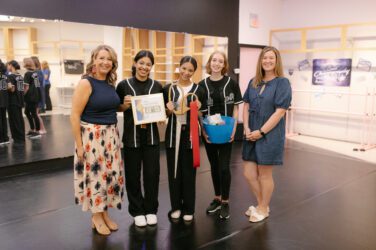 Four smiling girls in matching black outfits hold certificates and gifts, standing between two women at Synergy Dance Project’s studio, with mirrors, ballet barres, and posters visible in the background.