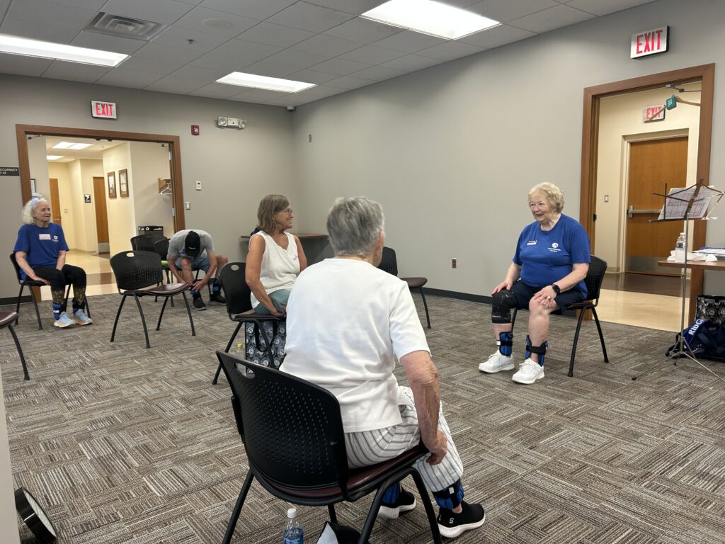 Adults sit in chairs spaced out in semi-circle rows, wearing leg weights.