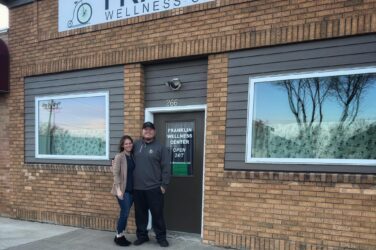 Two people stand smiling in front of the Franklin Wellness Center, a brick building with large windows. The entrance sign reads “Open 24/7,” and a poster advertises Well Made Meal Prep services for visitors.