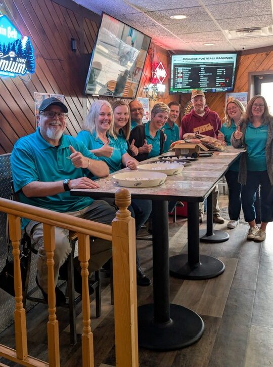 Montevideo Area Community Foundation Advisory Board Member sporting matching teal polo shirts pose for a group photo at their meat raffle.