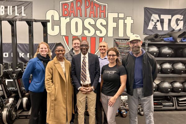 Sen. Bobby Joe Champion and business owner Lourdez Schwab with a group of people standing inside a gym in front of a large wall sign that reads “Bar Path CrossFit.”