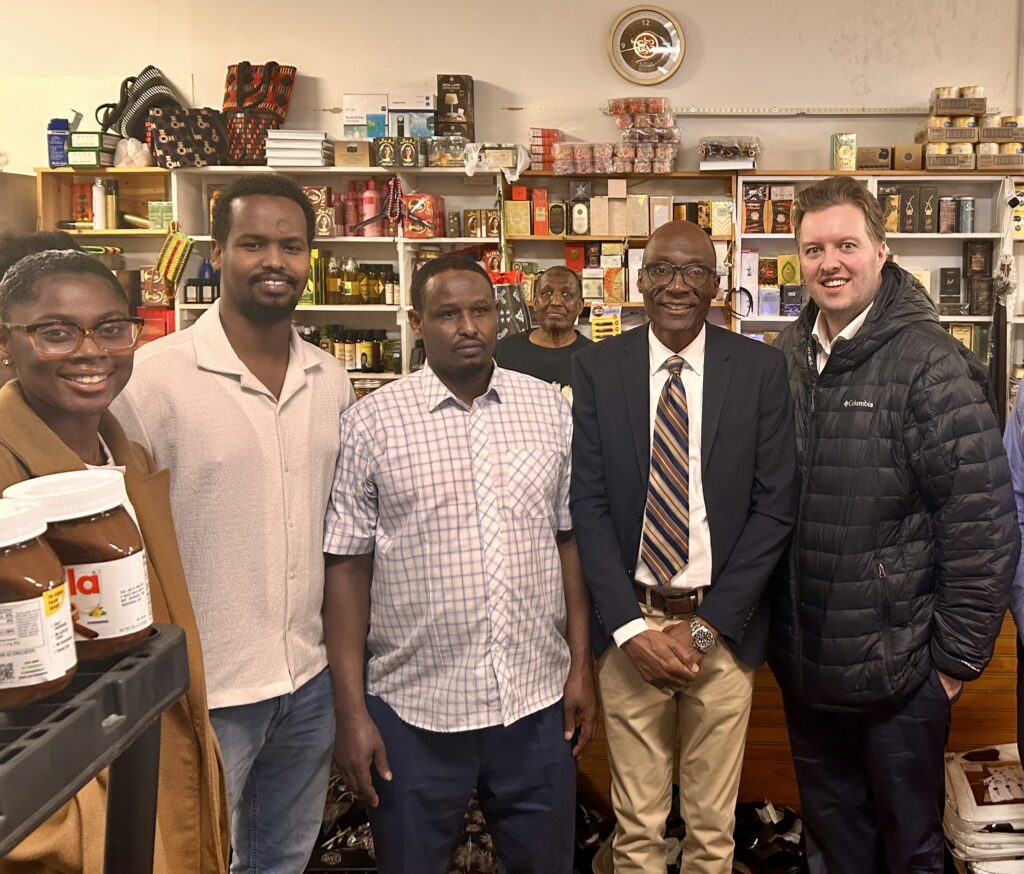 Sen. Bobby Joe Champion and a group of people standing inside Mubarak Food & Grocery with owner Luqman Mohamed. Shelves in the background are filled with packaged food items, jars, and bottles.
