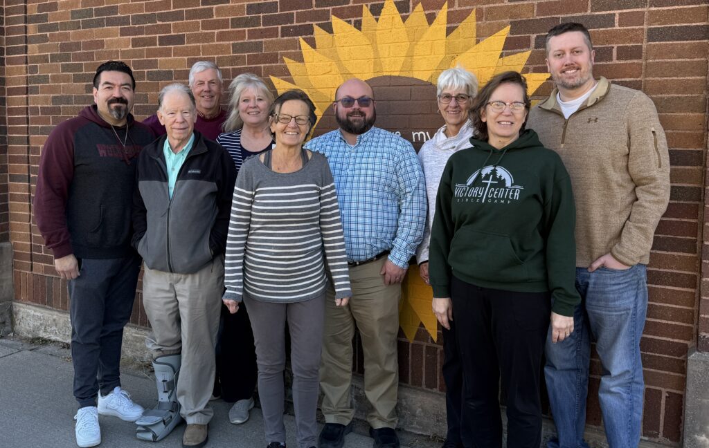 Nine adults stand smiling in front of a brick wall with a large yellow sunflower mural. They are dressed casually, and one person has a walking boot on their left foot.